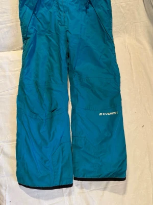 Blå täckbyxor från Everest stl 134 - Blå täckbyxor från Everest i storlek 134. Byxorna har raka ben, förstärkta knän och praktiska fickor. Tillverkade i slitstarkt syntetmaterial med mjukt foder på insidan. Perfekta för kalla dagar och vinteraktiviteter.