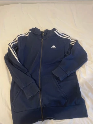 Mörkblå Adidas hoodie med dragkedja - Säljer en mörkblå kofta  från Adidas med klassiska vita ränder längs ärmarna och Adidas-logga på bröstet. Tröjan har huva, långa ärmar, två fickor fram och hel dragkedja. Perfekt för chill eller träning, riktigt skön och sportig stil.