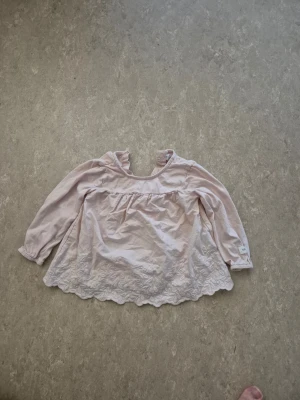 Blus - Säljer en söt ljusrosa blus från Newbie i storlek 74. Blusen har långärmade ärmar med resår, hals med knapp och fina broderade spetsdetaljer längs nederkanten. Perfekt för dig som gillar romantisk och mjuk stil.