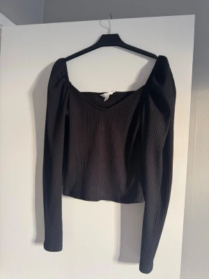 Svart ribbad topp med puffärm - Säljer en svart ribbad topp i storlek M med lång puffärm och snygg v-ringad hals. Toppen har en croppad passform och är tillverkad i mjukt, stretchigt material som sitter bekvämt. Perfekt att styla med jeans eller kjol för en cool look.