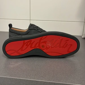 Grå sneakers från Christian Louboutin - Snygga grå sneakers i mocka från Christian Louboutin med den ikoniska röda sulan. Skorna har snörning och en stilren design som passar perfekt för en trendig look.