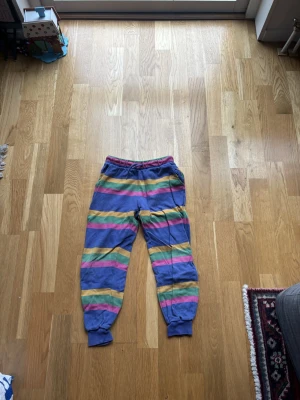 Randiga mjukisbyxor från Mini Rodini - Färgglada mjukisbyxor från Mini Rodini med breda ränder i blått, gult, rosa och grönt. Byxorna har resår och snörning i midjan samt muddar vid bensluten. Tillverkade i mjuk bomull som är skön att ha på sig. Perfekt för chill dagar.