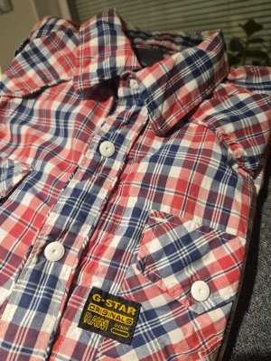 Rutig skjorta från G-Star RAW XL - Säljer en rutig skjorta från G-Star RAW i storlek XL. Skjortan har ett klassiskt rutmönster i rött, blått och vitt, två bröstfickor med knappar och tydliga vita knappar framtill. Materialet är mjuk bomull och skjortan har lång ärm och krage.