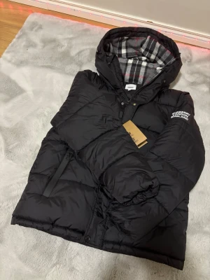 Svart pufferjacka från Burberry - Svart pufferjacka från Burberry med huva och klassiskt rutigt foder. Jackan har dragkedja framtill, sidofickor med dragkedja och Burberry London England-logga på ärmen. Materialet är polyester och jackan har en quiltad design som ger extra volym och värme.