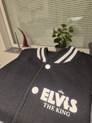Svart och grå bomberjacka Elvis - Snygg bomberjacka med svart kropp och grå ärmar, vita tryckknappar och coolt Elvis-tryck fram och bak. Jackan har ribbade muddar och krage med vita ränder, samt två fickor med vit kant. Perfekt för dig som gillar retro och streetstyle.