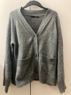 Grå stickad kofta  - Mysig grå stickad kofta från 157 i storlek M. Koftan har v-ringad hals, stora ribbade fickor framtill och knäpps med tre knappar. Perfekt för lager-på-lager och chill dagar. Materialet är mjukt och ger en skön känsla.