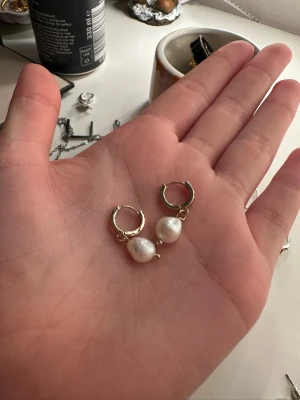Guldiga örhängen med pärlor - Säljer ett par snygga örhängen med guldfärgade ringar och droppformade vita pärlor. Perfekt accessoar för att lyfta din stil, de har en enkel men elegant design som passar till många looks.