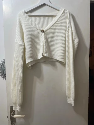 Vit croppad kofta med knappar - Säljer en vit croppad kofta med ribbad struktur och två stora knappar framtill. Koftan har v-ringad hals och långa ärmar med breda muddar. Perfekt för att ge din outfit en fräsch och trendig touch.