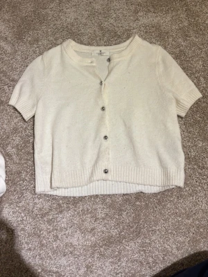 Vit kortärmad kofta från H&M - Säljer en vit, kortärmad kofta från H&M med rund hals och knappar framtill. Koftan har ribbade muddar vid ärmslut och nederkant, vilket ger en snygg och enkel look. Perfekt att slänga över en topp för extra stilpoäng.