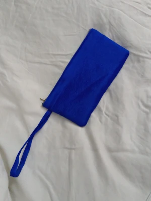 Blå kuvertväska till mobilen - Snygg blå kuvertväska med praktisk dragkedja och handledsrem. Väskan är tillverkad i ett slätt tygmaterial och har ett rymligt fack med plats för kort och småsaker. Perfekt att ha med sig när du vill bära det viktigaste. 