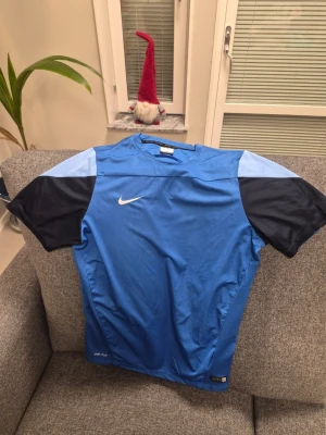 Nike Dri-Fit blå fotbollströja L - Nike Dri-Fit fotbollströja i storlek L med autentisk team-märke. Tröjan är blå med mörkblå och ljusblå detaljer på ärmarna, tillverkad i lätt och ventilerande polyester som håller dig torr under träning eller match. Snygg och sportig design med klassisk Nike-logga på bröstet.