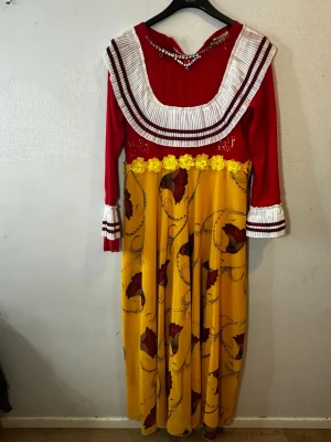 Färgstark långklänning från B&R Fashion - Säljer en unik långklänning med röd överdel och plisserad vit krage med röda ränder. Kjolen är gul med blommigt mönster och dekorativa gula blommor i midjan. Långärmad med plisserade manschetter. Perfekt för dig som gillar färg och detaljer!