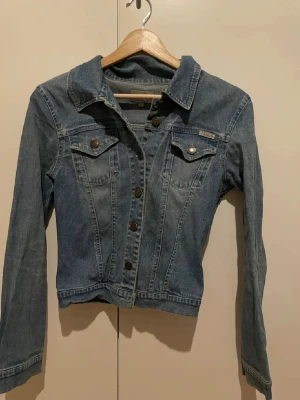 Blå jeansjacka från Tessie - Snygg blå jeansjacka från Tessie med klassisk krage, långa ärmar och bröstfickor med knappar. Jackan har en rak passform och är dekorerad med metallknappar där Tessie är ingraverat. Perfekt att slänga över en hoodie eller t-shirt.