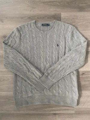 Grå kabelstickad tröja Polo Ralph Lauren - Kabelstickad grå tröja från Polo Ralph Lauren i storlek M. Klassisk rund halsringning, ribbade muddar och ikonisk broderad logga på bröstet. Perfekt till höst och vinter för en clean och tidlös look. Mycket bra skick, passar till allt😁