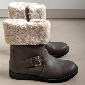 Bruna boots med fuskpäls från H&M - Säljer ett par bruna boots från H&M i storlek 36. De har en rund tå, platt sula och är fodrade med mjuk beige fuskpäls upptill. Bootsens ovandel är i syntetmaterial och de har en snygg spänndetalj på sidan samt dragkedja för enkel påtagning.