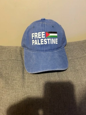 Blå keps med trycket Free Palestine  - Blå keps med böjd skärm och justerbart spänne bak. Framsidan har ett tryck med texten 'FREE PALESTINE' och en flagga i rött, grönt, vitt och svart. Kepsen är tillverkad i bomull och har en klassisk, avslappnad look som passar till streetwear.FREE PALESTINE KEPS - GÅRDA WASHED  DEN ÄR HELT Blå MEN SISTA TVÅ BILDRER BARA VISAR HUR DEN SER UT. Detaljinformation:  Tillverkad av 100 procent bomull One Size Justerbar vid kepsens baksida Gjord av: 100% bomull  