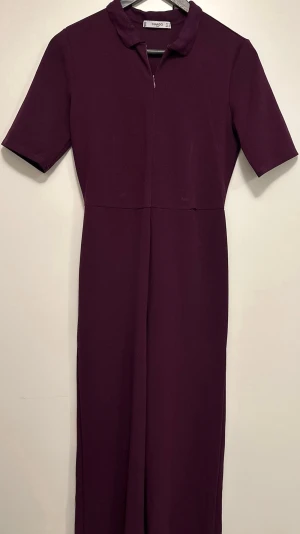Lila byxdress från Mango - Säljer en stilren lila byxdress från Mango i storlek S. Modellen har korta ärmar, krage och dragkedja framtill. Materialet är mjukt och har en snygg, slät finish.