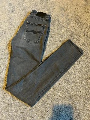 Mörkgrå jeans från Nudie Jeans - Säljer ett par mörkgrå jeans från Nudie Jeans med klassisk femficksdesign och snygga kontrastsömmar. Jeansen har rak passform och är tillverkade i slitstarkt bomullsmaterial. Bakfickan har Nudie Jeans logga och en cool broderad detalj.