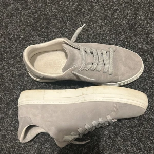 Gråa sneakers i mocka från Selected Homme - Snygga grå sneakers i mocka från Selected Homme. Skorna har rund tå, platt vit sula och grå snörning. Pris kan diskuteras. Skickar ofta samma dag! Box finns med.