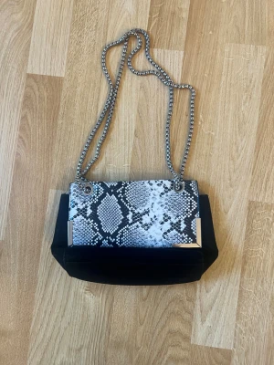 Svart axelväska med ormmönster - Snygg svart axelväska med lock i ormmönstrad design och silverfärgade metalldetaljer. Väskan har en kedja som axelrem och ett rymligt fack med dragkedja på insidan. Perfekt för dig som gillar att sticka ut med coola detaljer.