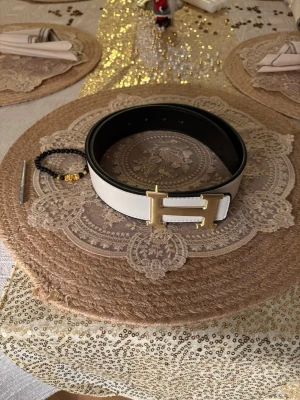 Vit bälte - Säljer en stilren vit väst med bred passform och en stor H-formad guldfärgad spänne framtill. Västen är tillverkad i skinn och har en modern look som sticker ut. Perfekt för dig som vill ha något extra till outfiten.