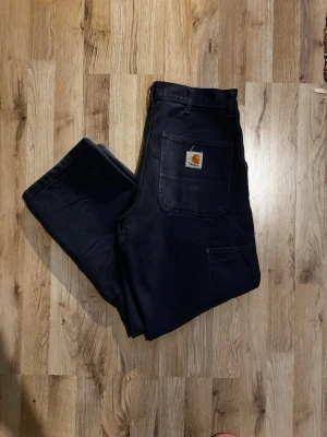 Blåa cargopants från Carhartt - Blåa cargopants från Carhartt med raka ben och flera praktiska fickor, inklusive bakfickor med Carhartt-logga. Byxorna har normal passform och är tillverkade i slitstark bomull. Perfekta för dig som gillar streetwear och funktionella detaljer.