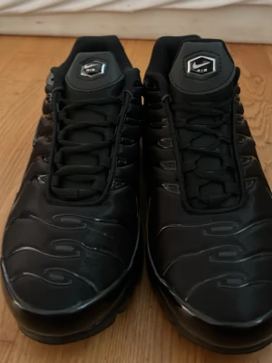 Nike Air Max Plus svarta sneakers - Nike Air Max Plus sneakers i helsvart med glänsande detaljer och synlig Air-enhet i sulan. Snyggt vågigt mönster på ovandelen, snörning och Tn-logga bak. Skorna har en sportig och futuristisk vibe, perfekt för dig som gillar streetstyle.