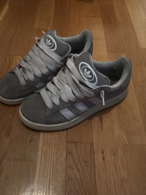 Grå Adidas Campus sneakers mocka - Säljer ett par grå Adidas Campus sneakers i mocka med klassiska vita ränder på sidorna och breda vita skosnören. Låg modell med rund tå och vit sula. Adidas-logga på plösen och hälen. Perfekta för dig som gillar retrovibbar och streetstyle.