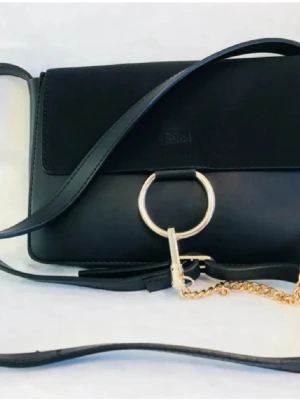 Svart axelväska från Chloé - Säljer en stilren svart axelväska från Chloé med snygga gulddetaljer och en stor metallring framtill. Väskan har en lång axelrem och en kedja som ger extra edge. Perfekt för dig som gillar minimalistisk design och lyxiga accessoarer.