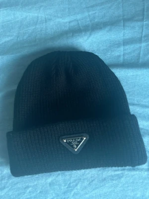 Svart stickad mössa från Prada - Snygg svart stickad mössa från Prada med klassisk uppvikt kant och metallmärke framtill. Mössan har en enkel och stilren design som passar perfekt till kalla dagar. Märket är i form av en triangel och ger en exklusiv touch.
