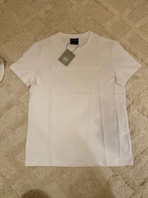 Vit t-shirt från Canada Goose - Canada Goose Tröja – Vit (Helt ny) 👕 Storlek: M 🤍 Färg: Vit ✨ Skick: 10/10 – helt ny, aldrig använd 🏷️ Detalj: Originaltag kvar 💎 Märke: Canada Goose