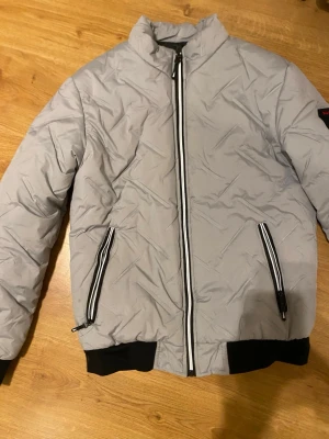 Grå pufferjacka med Fashion G-märke - Snygg grå pufferjacka med svart och vit dragkedja framtill och på fickorna. Jackan har ett coolt Fashion G-märke på ärmen och svarta muddar vid nederkant. Perfekt för dig som gillar streetstyle och vill hålla dig varm under kyliga dagar.