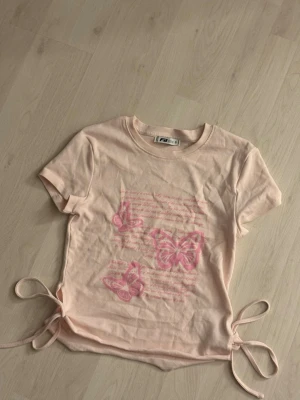 Ljusrosa t-shirt med fjärilar FB Sister - Söt ljusrosa t-shirt från FB Sister i storlek S. Framsidan har tryck med rosa fjärilar och text, samt snörning i båda sidorna för en extra cool detalj. T-shirten är kortärmad och har en rund hals. Perfekt för dig som gillar gulliga prints och mjuka färger.