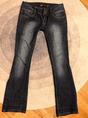 Mörkblå bootcut jeans från JSFN - Snygga mörkblå jeans från JSFN med bootcut-snitt och låg midja. Jeansen har klassiska fem fickor, dekorativa sömmar och slitningar framtill. Materialet är jeans med kontrastfärgade sömmar och flera knappar i midjan. Perfekt för dig som gillar en avslappnad stil.  SÄLJER för att de är för små