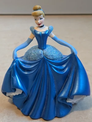  Askungen med blå balklänning med puffärmar - Säljer Askungen med ikonisk blå balklänning. Hela figuren och balklänningen är av plast  Askungen från Disney. Klänningen har glittrigt liv och kjol, puffärmar och markerad midja. Lång modell med vackra draperingar och axelbandslös look. Perfekt för dig som älskar sagofigurer.