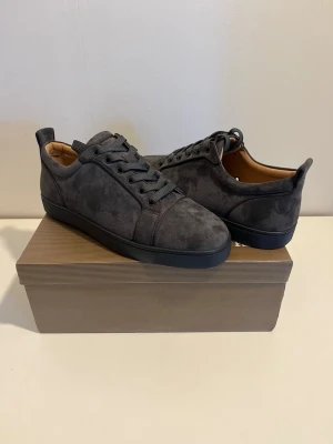 Christian Louboutin mörkgrå - Snygga svarta sneakers från Christian Louboutin i mocka med klassisk röd sula och svarta snören. Insidan är i beige skinn och yttersulan har Louboutin-loggan i rött. Modellen heter Lou Spikes Oralto CLF/GLITT och har en stilren, rund tå.