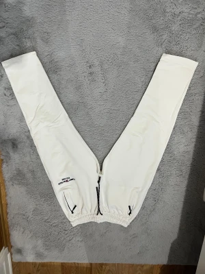 Vita mjukisbyxor från Tommy Hilfiger - Säljer ett par vita mjukisbyxor från Tommy Hilfiger med broderad logga på ena benet och svarta detaljer vid fickorna. Byxorna har elastisk midja med snörning och raka ben. Perfekta för chill eller träning.