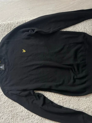 Svart stickad tröja från Lyle & Scott - Svart stickad tröja från Lyle & Scott med klassisk gul broderad fågel på bröstet. Tröjan har rund hals, ribbade muddar och lång ärm. Perfekt för dig som gillar stilrena och enkla plagg med en liten detalj.