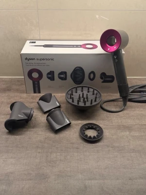 Dyson Supersonic hårfön med tillbehör - Dyson Supersonic hårfön i metallicgrå med en snygg rosa front. Medföljer flera svarta munstycken och en diffuser för olika stylingmöjligheter. Modern design och kraftfull motor, perfekt för att snabbt torka och styla håret. Materialet är plast och metall.