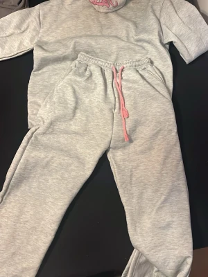 Grå mjukisbyxor med rosa tryck - Säljer ett par grå mjukisbyxor från NA-KD med rosa snörning i midjan och stort rosa tryck på ena benet. Byxorna har elastisk midja och är gjorda i mjukt bomullsmaterial, perfekta för chill dagar. Passar dig som gillar streetstyle och comfy vibes.