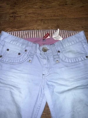 Lågmidjade ljusblå jeansbyxor  - Säljer ett par ljusblå jeansbyxor från Divided med klassisk femficksdesign och snygga nitar vid fickorna. Insidan av midjan har ett rosa och grått randigt tyg, och bak finns en rosa patch med ros-tryck. Lågmidjade. DET STÅR 40/L MEN DET PASSAR XS/S
