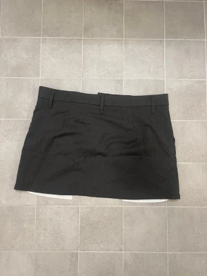 Svart kortkjol från H&M Divided - Säljer en svart kortkjol från H&M Divided i storlek M. Kjolen har bälteshällor, dragkedja och knapp framtill samt diskreta veck för en snygg passform. Materialet är polyester och viskos, vilket gör den mjuk och bekväm. Perfekt för dig som gillar stilrena plagg.