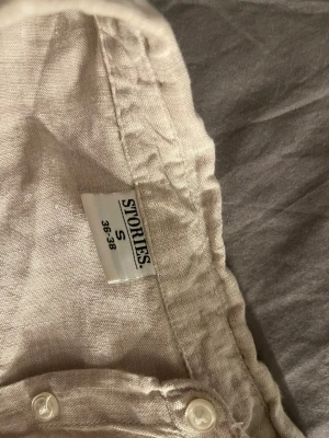 Beige linneskjorta från Stories S - Säljer en beige skjorta från Stories i storlek S. Skjortan är tillverkad i linne och har klassisk krage, knappar framtill och långa ärmar. Perfekt för dig som gillar en chill och stilren look med naturliga material.