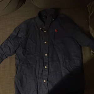 Mörkblå  linneskjorta från Ralph Lauren - Snygg mörkblå skjorta från Ralph Lauren i storlek XS. Klassisk design med knappar hela vägen och lång ärm. Skjortan har en röd broderad logga på bröstet och är gjord i mjukt linne. Perfekt för dig som gillar stilrena och tidlösa plagg.