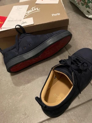 Svarta sneakers från Christian Louboutin - Säljer ett par svarta sneakers från Christian Louboutin med klassisk röd sula. Skorna har rund tå, snörning och är tillverkade i mocka med beige innersula. Perfekt för dig som vill ha en lyxig och stilren look.