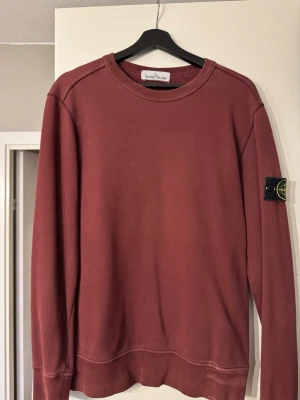 Mörkröd sweatshirt från Stone Island - Säljer en mörkröd sweatshirt från Stone Island med klassisk rund hals och ribbade muddar. Tröjan har lång ärm och den ikoniska Stone Island-loggan på vänster ärm. Materialet är mjuk bomull och passformen är normal. Perfekt för dig som gillar streetwear och snygga detaljer.