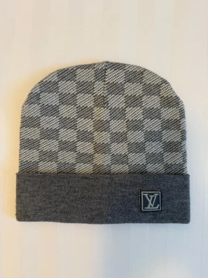 Gråvit ruttig Luice vitton vest - Säljer en stilren väst från Louis Vuitton i grått och vitt med rutigt mönster. Västen har en bred grå kant nedtill och ett broderat LV-emblem framtill. Materialet är stickat och känns mjukt mot huden. Perfekt för dig som gillar exklusiva märken och klassisk design.