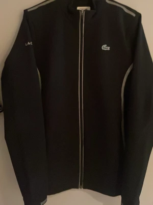 Svart Lacoste Sport vindjacka Novak Djokovic - Säljer en svart Lacoste Sport Novak Djokovic vindjacka i storlek M. Jackan har reflekterande detaljer längs dragkedjan, ärmar och axlar samt klassisk Lacoste-logga på bröstet. Tillverkad i polyester och har en clean, sportig look med hög krage och långa ärmar.