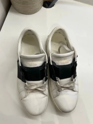 Valentino opens - Säljer ett par vita Valentino Garavani sneakers med bred svart rem över snörningen. Skorna har rund tå, platt sula och klassisk låg profil. Tillverkade i skinn med vita snören och diskret logga i guld på plösen. Perfekta för dig som gillar stilrena designer.