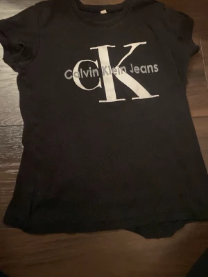 Svart Calvin Klein Jeans t-shirt - Svart t-shirt från Calvin Klein Jeans med stort CK-tryck i vitt på bröstet. Klassisk rund hals och korta ärmar. T-shirten är gjord i mjuk bomull och har en clean, stilren look som passar perfekt till jeans eller shorts. Väldigt liten i storleken så den passar xs.